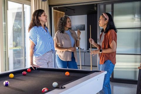Diverse women planning pool shot at black felt table in modern sunlit living room, social