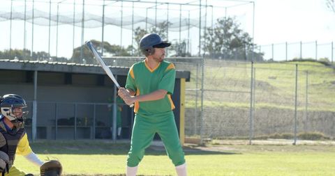 Amateur baseball batter at plate wearing green uniform preparing to swing on sunny field