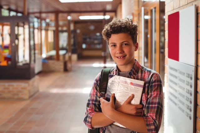 Teenage boy standing in a school corridor, holding a notebook and smiling. Ideal for educational content, school-related advertisements, and youth-focused campaigns. Can be used to depict student life, academic environments, and back-to-school promotions.