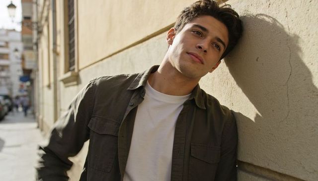 Young man leaning on sunlit stucco wall watching urban alley with thoughtful expression