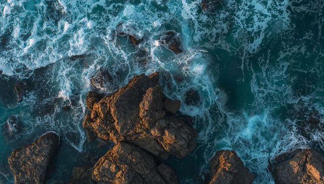 Sunlit rocky outcrop with teal surf crashing around jagged coastline, aerial drone seascape