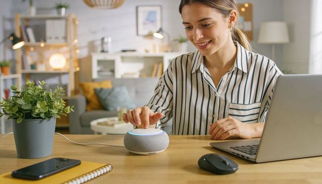Woman tapping smart speaker at home workspace while using laptop, cozy living room office