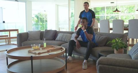 Friends watching casual game on couch drinking soda, eating pizza in bright living room