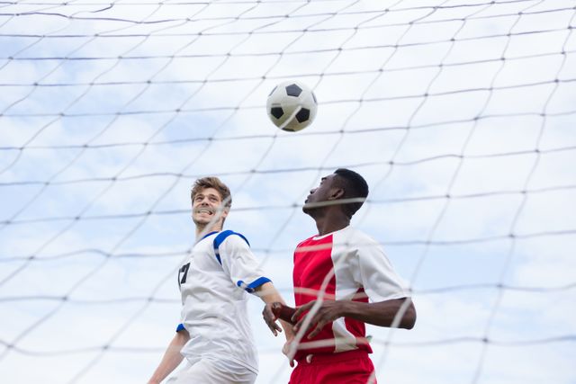 Two male soccer players in action near the goal, competing for the ball. One player is wearing a white uniform while the other is in a red uniform. The sky is clear, indicating a sunny day. This image can be used for sports-related content, teamwork concepts, athletic promotions, or articles about soccer matches.
