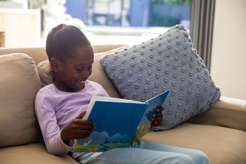 African american girl reading picture book on sofa in sunlit living room, cozy moment