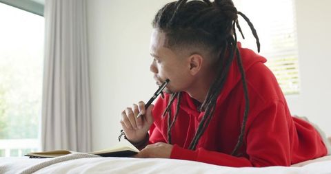 African american man journaling on bed wearing red hoodie, contemplating ideas in daylight