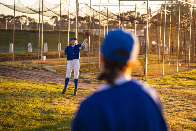 Female baseball players warming up on a field during sunset. One player is catching a ball while the other looks on. Ideal for use in sports training materials, youth sports promotions, teamwork and athleticism concepts, and advertisements for sports equipment or apparel.