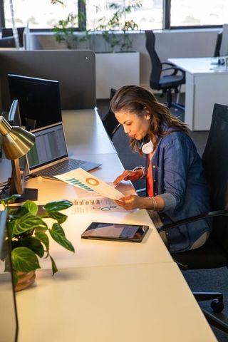 Reviewing business charts at modern open-plan desk with laptop, tablet, headphones and plants