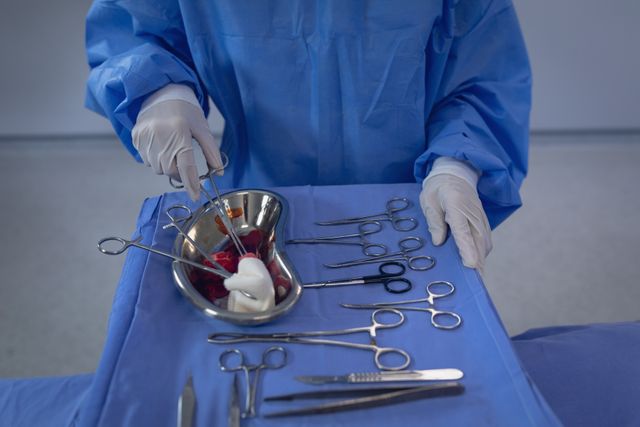 Surgeon wearing blue scrubs and gloves is handling surgical scissors over a tray of surgical instruments in an operating room. Ideal for use in medical publications, healthcare promotions, and educational materials related to surgery and medical procedures.