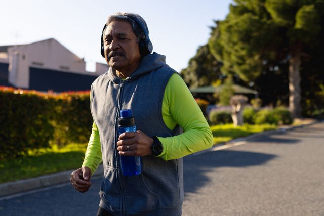 Happy senior biracial man jogging on street wearing headphones and holding water, with copy space. Retirement, active lifestyle and senior health concept.