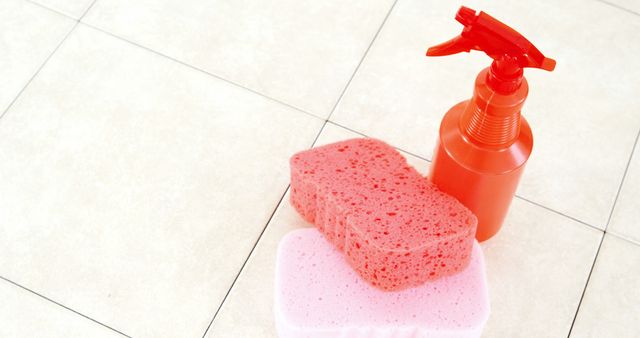 A red spray bottle and pink sponges are placed on a tiled floor, with copy space. Cleaning supplies like these are essential for maintaining hygiene and cleanliness in homes and workplaces.
