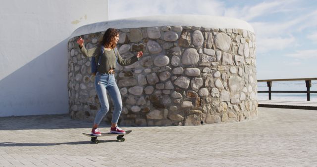 Young woman enjoying skateboarding beside a stone wall near ocean. Ideal for concepts of youth lifestyle, outdoor activities, health, and fun. Suitable for advertising campaigns, social media, and articles related to skateboarding and healthy living.