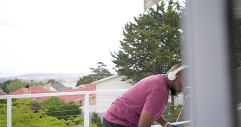 Contemplative african man leaning on balcony railing wearing headphones overlooking suburb