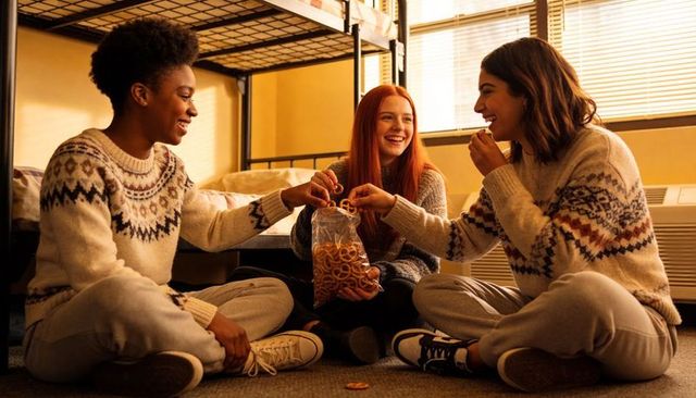 College roommates sharing snacks on dorm floor in cozy knit sweaters, laughing and chatting
