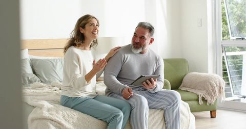 Middle-aged couple sharing tender morning moment on bed, man holding tablet