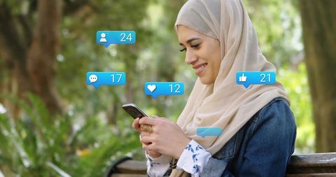 Smiling woman wearing hijab using smartphone on bench with social media notifications