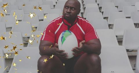 Focused rugby player sitting in empty stadium holding ball with gold star sparkles overlay