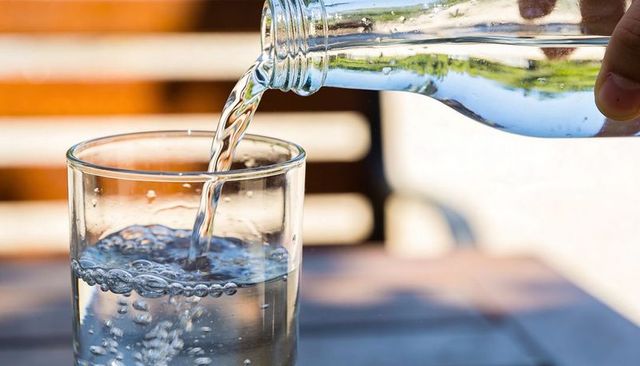 Refreshing water pouring from glass bottle into tumbler with bubbles on sunlit patio