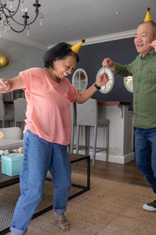 Happy senior couple dancing in living room wearing party hats and laughing over gifts