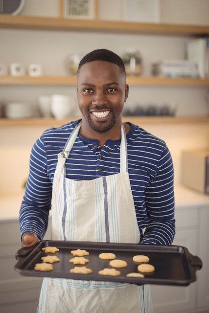 This image depicts a cheerful man holding a tray of freshly baked cookies in a home kitchen. He is wearing a striped shirt and an apron, suggesting he is engaged in baking. The bright and modern kitchen setting adds a warm and inviting atmosphere. This image can be used for promoting home baking, cooking classes, lifestyle blogs, or advertisements for kitchenware and baking products.
