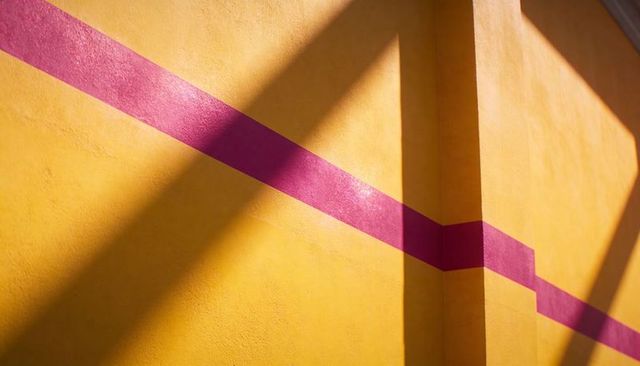 Yellow plaster wall with magenta diagonal stripe and column casting sharp geometric shadows