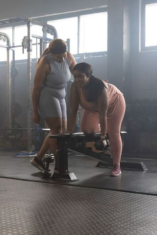 African american women training dumbbell row on bench with spotter, plus-size workout