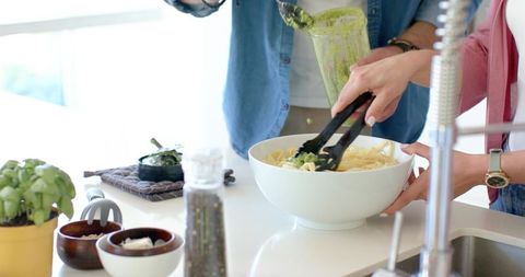 Couple tossing pesto pasta together in bright modern kitchen, casual home cooking