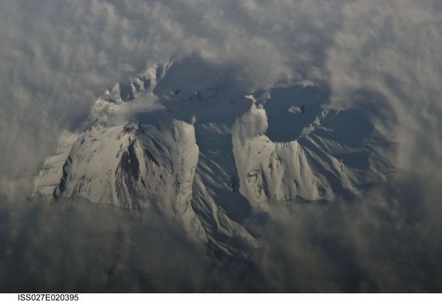 Aerial view of snow-covered Avachinsky Volcano with clouds surrounding it in the Kamchatka Peninsula, Russia. Kozelsky Volcano's breached crater is also visible. The image was taken from the International Space Station. Ideal for use in educational materials about geology, volcanic activity, and Earth observation, travel brochures highlighting natural landscapes, and environmental conservation campaigns.