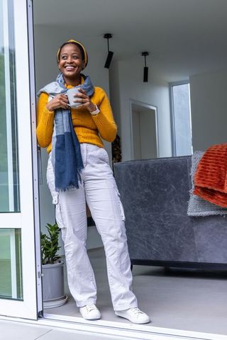 African american woman smiling and holding mug by sliding door in mustard sweater