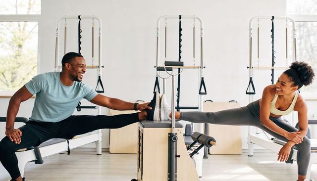 Fitness partners stretching on pilates reformers, smiling during assisted leg flexibility workout