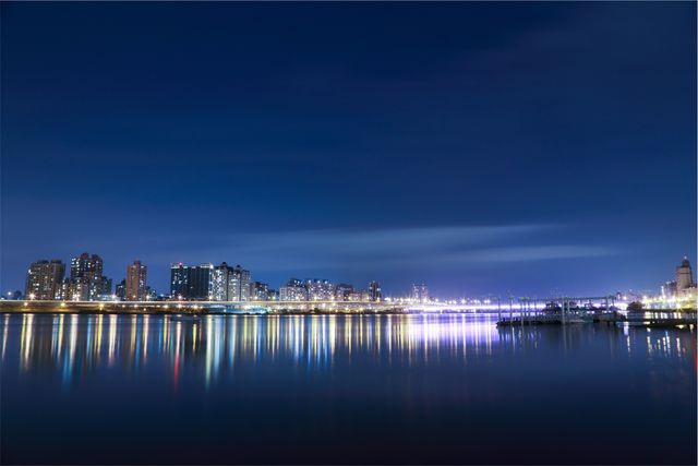 Panoramic view of city skyline during nighttime with bright lights reflecting on still water. Ideal for backgrounds, travel blogs, and websites focused on urban environments, city life, or architecture. Effective for use in promotions related to real estate, tourism, and city events.
