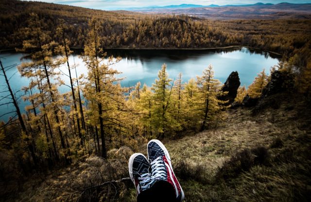 Person's feet in sneakers seen relaxing on a clifftop overlooking a serene lake and forest during autumn season. Vibrant foliage and distant mountains enhance the breathtaking scenery. Ideal for themes of travel inspiration, exploration, outdoor activities, tranquility, and nature appreciation.
