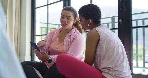 Two african american women sharing fitness app on phone wearing smartwatch on balcony