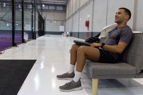 Young athlete relaxing on bench at indoor courts holding racket case and towel
