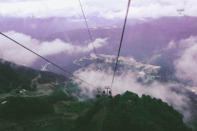 Cable cars are ascending over a high-altitude scenic valley surrounded by lush green mountains and partially covered by clouds. Ideal for promoting travel destinations, outdoor adventure content, and tourism websites. This conveys a sense of thrill and exploration in nature.