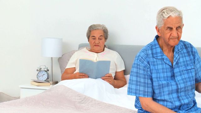 Elderly man and woman sharing peaceful moment in their bedroom; elderly woman reading book, elderly man looking thoughtful in pajamas. This video is ideal for topics related to senior living, retirement, homely atmosphere, and healthy daily routines.