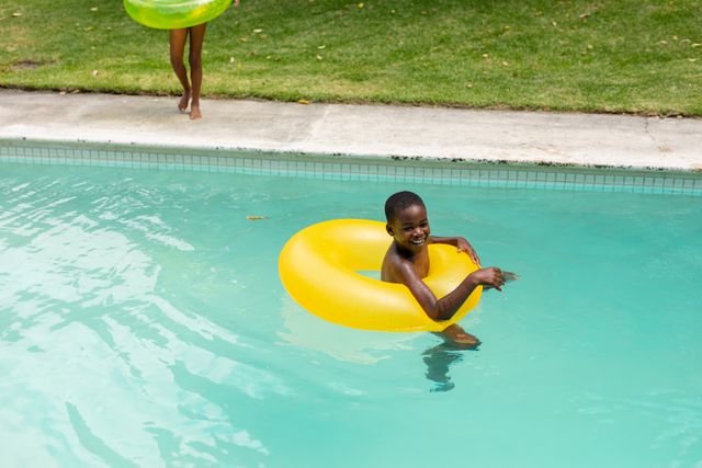 Young African American boy enjoying a sunny day in the pool with a bright yellow inflatable ring. Perfect for use in advertisements for summer activities, family vacations, children's leisure products, or outdoor fun. Highlights themes of joy, playfulness, and childhood.