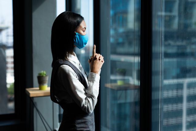 Asian woman wearing face mask standing and looking through window in an office. Ideal for use in articles or advertisements related to health and hygiene, workplace safety, pandemic precautions, or business environments during the COVID-19 pandemic.
