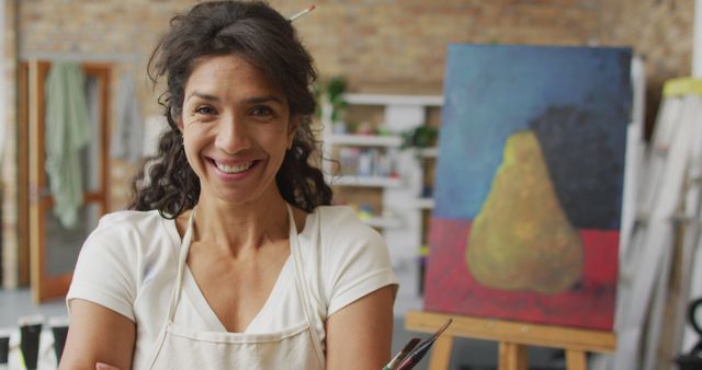 Female painter wearing an apron, smiling in front of her pear artwork in a creative studio. Great for art-related content, artist profiles, workshops, creative inspiration, artistic promotion.
