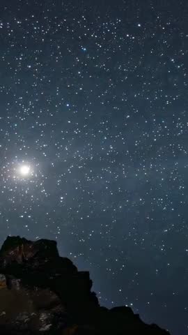 Vertical timelapse showing stars tracing celestial arc over rugged cliff with bright moon