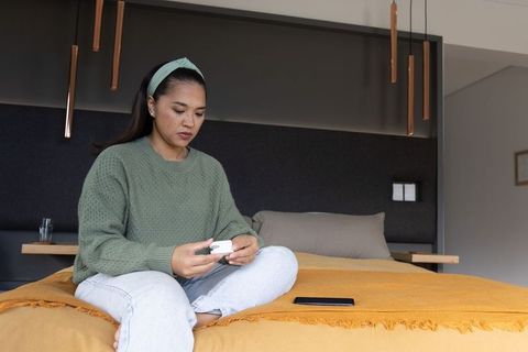 Mid-age asian woman sitting cross-legged on bed holding white device focusing on personal health