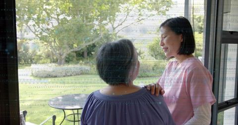 Caregiver daughter comforting elderly mother standing by patio door overlooking garden
