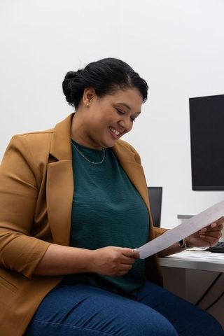 Asian professional woman smiling while reading document at modern office desk wearing tan blazer