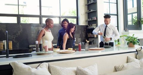Diverse friends gathering around kitchen island while man mixing cocktail in modern home