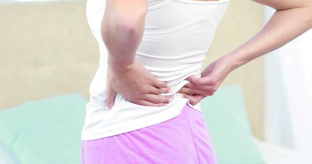 Woman in a white tank top and purple shorts holding her lower back, indicating pain or discomfort. This can be used in articles about back pain, muscle strain, healthcare, preventing back injuries, and general wellness.