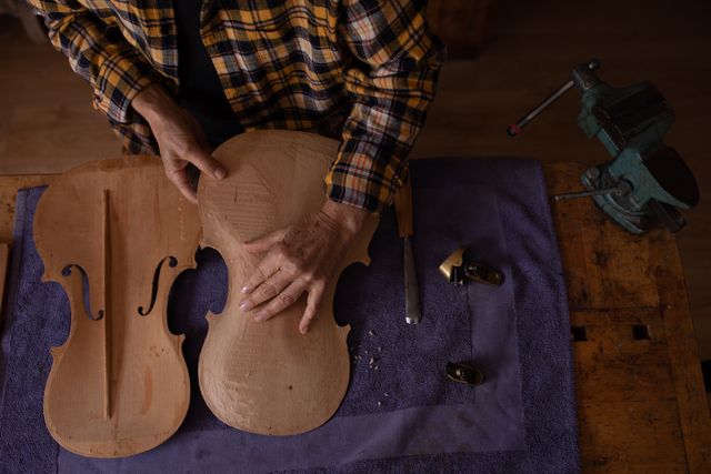 High angle view of a senior Caucasian female luthier standing and working on a violin at a workbench in her workshop, inspecting her handiwork and holding the unfinished violin body, with tools and a vise on the table.