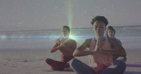 Three women practicing beach yoga at sunrise meditating in namaste pose with ocean horizon