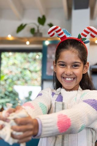 Smiling indian girl wearing candy-cane headband stretching dough while baking festive treats