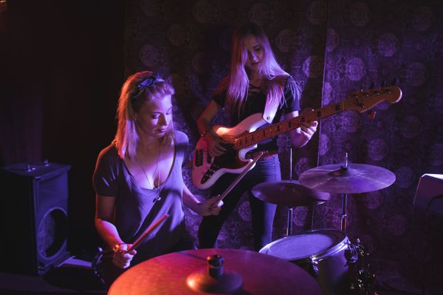 Two female musicians are performing live in a nightclub. The drummer is confidently playing the drums while the guitarist is focused on her instrument. This image can be used for promoting live music events, concerts, and nightlife entertainment. It is also suitable for articles or advertisements related to music, bands, and performing arts.