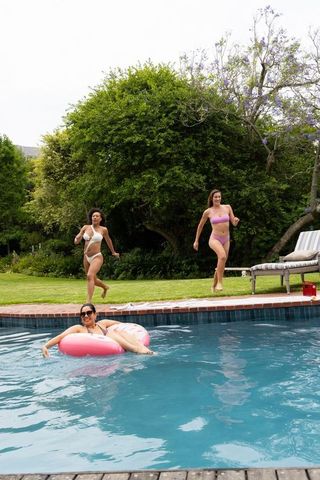 Women running toward pool while friend floats on pink donut inflatable in lush backyard